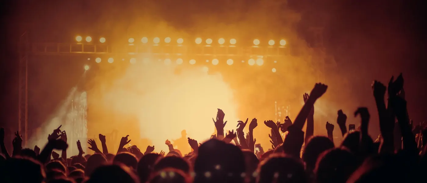 A concert crowd with raised hands in front of a stage filled with warm light and haze